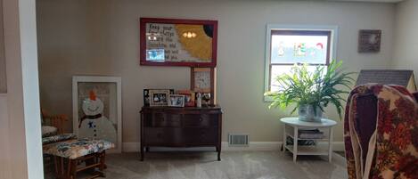 A cozy living room with a wooden dresser, a floral chair, a small table with a plant, a framed picture, and a wall clock.