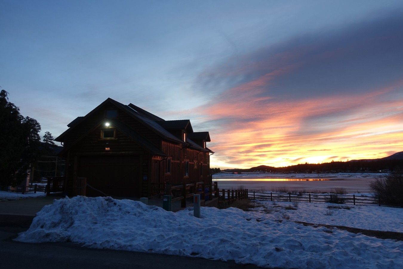 A wooden lodge with a lit window, a snow-covered ground, and a sunset over a frozen lake.