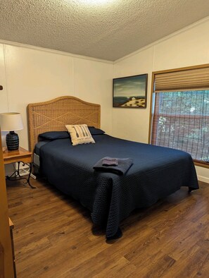 A bedroom with a bed, a nightstand, a lamp, a window with blinds, and a painting on the wall.