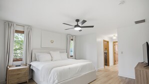 Un dormitorio con cama, mesita de noche, ventilador de techo y televisor sobre un soporte de madera.