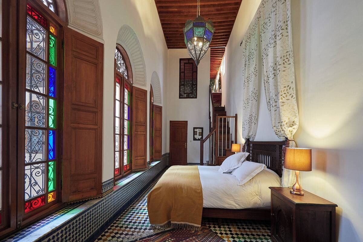 A bedroom with a bed, a nightstand, a lamp, a patterned rug, and a view of a staircase.