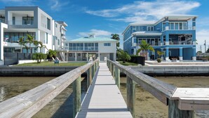 Una propiedad frente al agua con un paseo marítimo de madera que conduce a una casa moderna con techo azul, rodeada de palmeras y un cielo despejado.