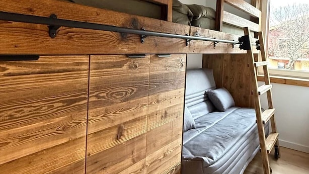 Ein Stockbett mit Holzkopfstütze, einer Leiter und einem Fenster mit Blick auf Bäume.