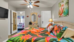 Quarto com cama, ventilador de teto, televisão, porta e parede com pintura.