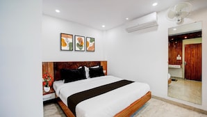 A hotel room with a bed, a nightstand with a vase of flowers, a wall-mounted air conditioner, and a mirror reflecting the bathroom.