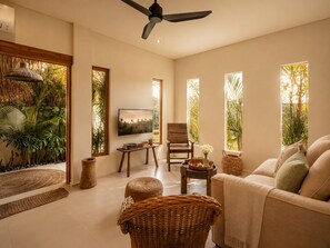 A living room with a ceiling fan, a TV, a sofa, a chair, a small table, and large windows with plants. - Rice Field Villa with Rooftop Jacuzzi (Canggu)