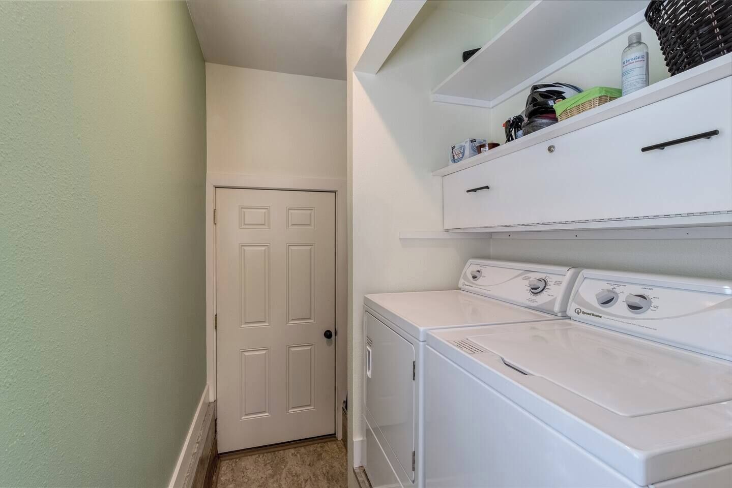 A laundry room with a white door, a washing machine, and a dryer. There are shelves above the dryer with various items.