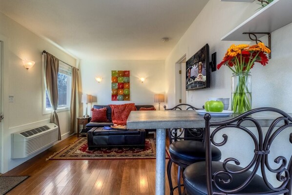 A cozy living room with a sofa, a dining table, and chairs. There is a window with curtains, a wall-mounted TV, and a vase with flowers.
