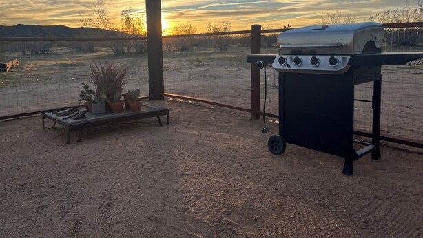 Ein Grill mit Schornstein, eine Bank mit Topfpflanzen und Blick auf ein eingezäuntes Gelände mit Sonnenuntergang im Hintergrund.