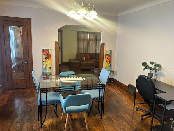 Uma sala de jantar com uma mesa de vidro, cadeiras azuis, chão de madeira, um sofá e um quadro.