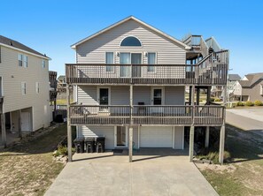 A two-story residential building with a front garage, a covered balcony on the second floor, and a small deck area. - Dollar Short: Semi-Oceanfront | Private Pool | Hot Tub (Kill Devil Hills)