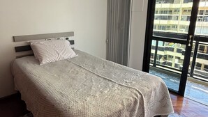 Ett sovrum med en säng, en luftkonditioneringsanläggning, ett fönster med gardiner och utsikt över byggnader utanför.
