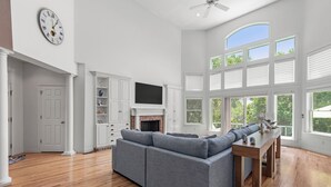 Un salón espacioso con un sofá seccional gris, una mesa de centro de madera y una chimenea. Se aprecia un ventanal grande con contraventanas blancas y un ventilador de techo.
