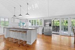 Cocina moderna con una isla central, taburetes de bar, iluminación colgante y vistas al exterior a través de amplios ventanales.