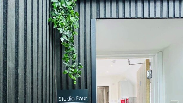 A modern interior with a sign reading "Studio Four" indicating a room or area. A vertical wooden paneling is visible on the left, and a hallway leads to a kitchen area with white cabinets and a wooden countertop. - Studio 4@Bransford Rd (Worcester)