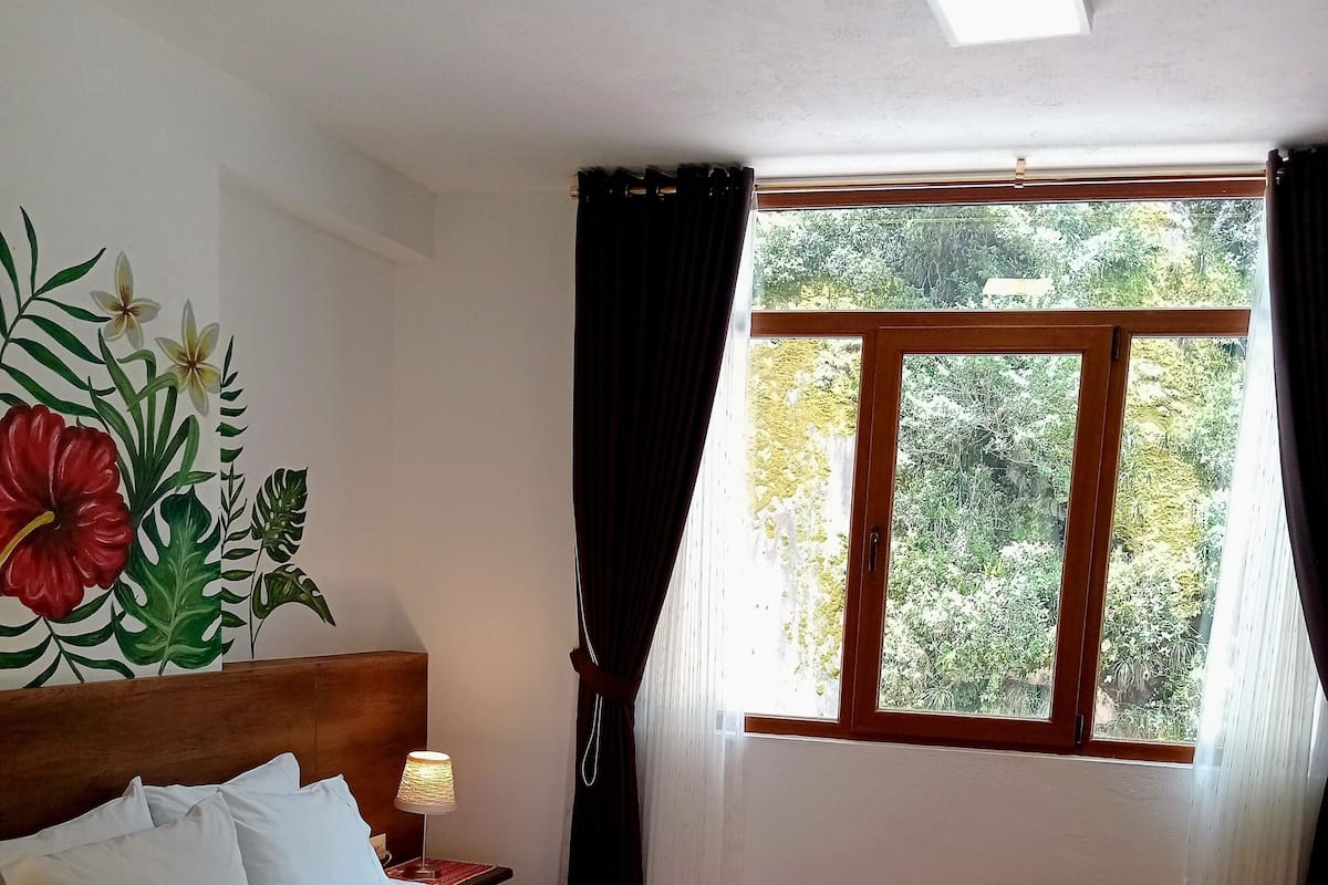 Ein ordentlich bezogenes Bett mit weißen Leinen und einer braunen Tagesdecke, ein Nachttisch mit Lampe und ein Fenster mit Vorhängen.