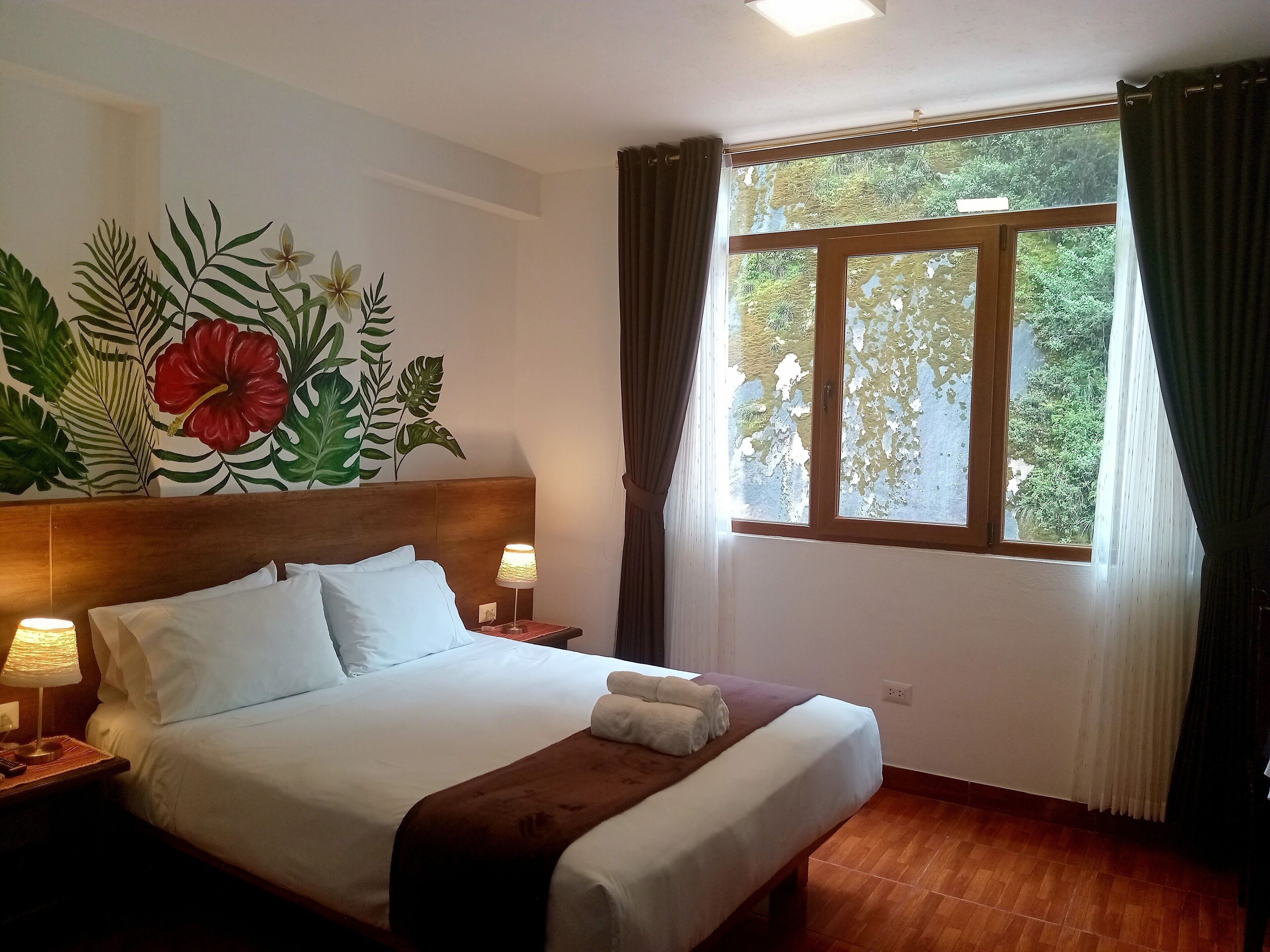 Ein ordentlich bezogenes Bett mit weißen Laken und einer braunen Tagesdecke, ein Nachttisch mit Lampe und ein großes Fenster mit Vorhängen.