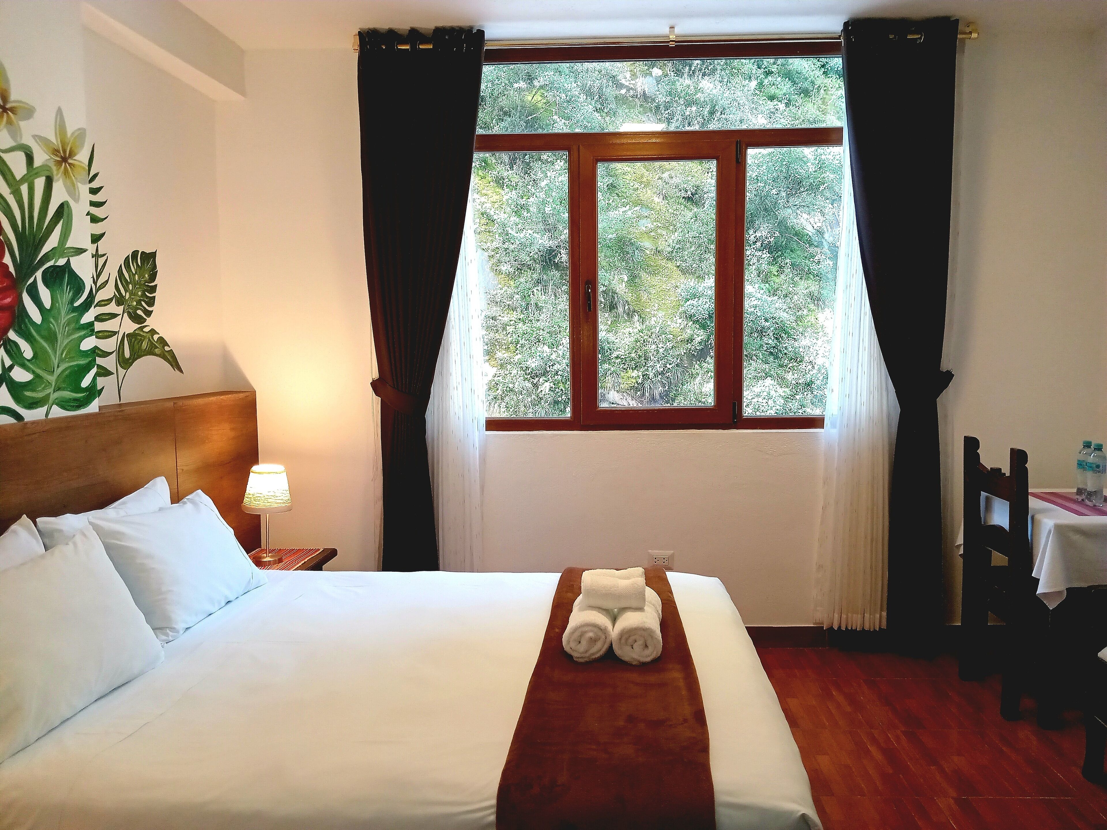 Une chambre d’hôtel avec un lit, une table de chevet avec une lampe, une fenêtre avec des rideaux et une vue sur la verdure.