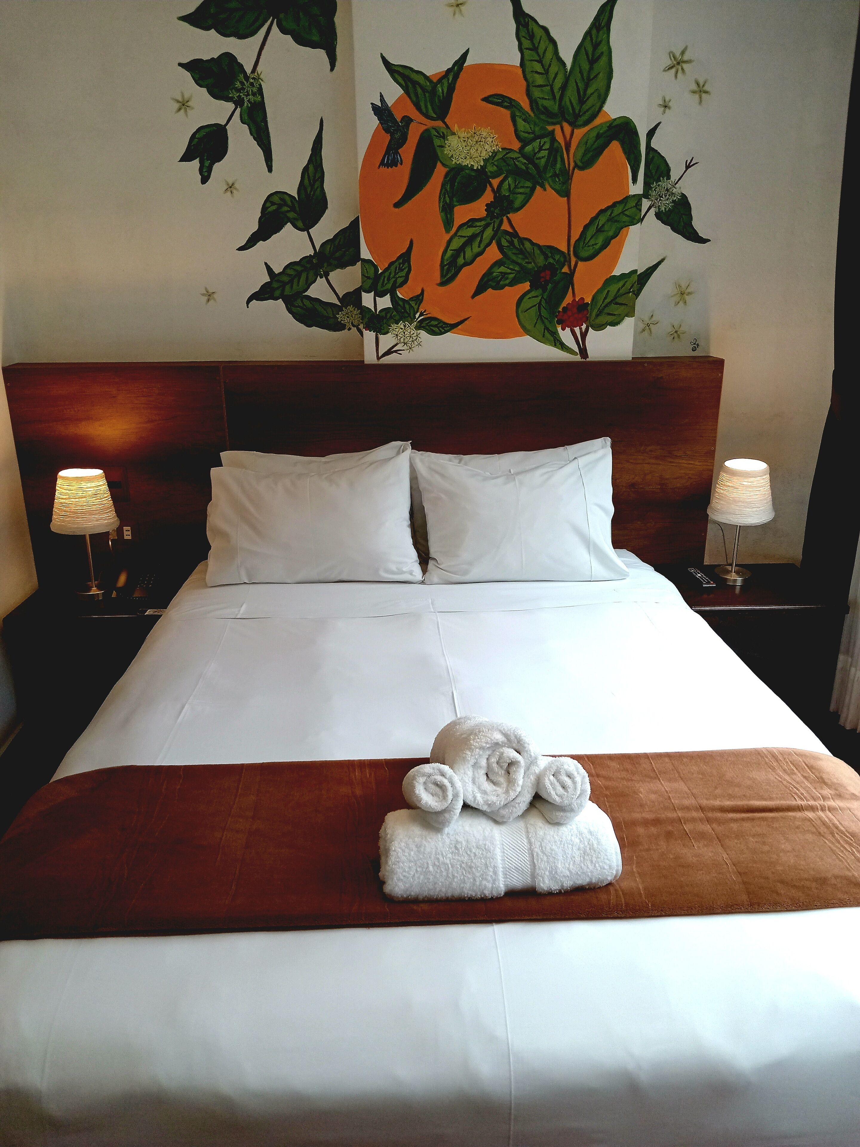 A neatly made bed with white linens, a brown throw blanket, and rolled towels at the foot of the bed. There are two bedside lamps on either side of the bed.