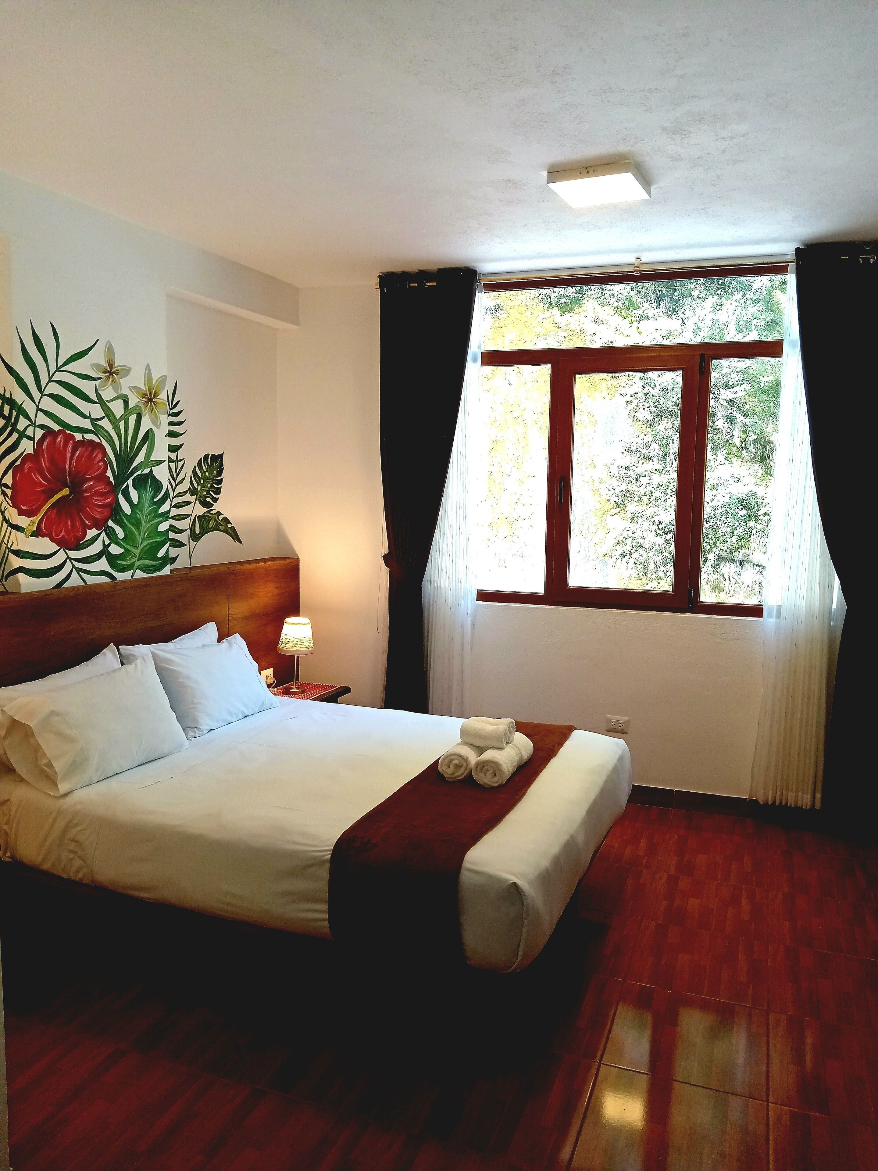 A hotel room with a bed, a nightstand, a window with curtains, and a wall mural.