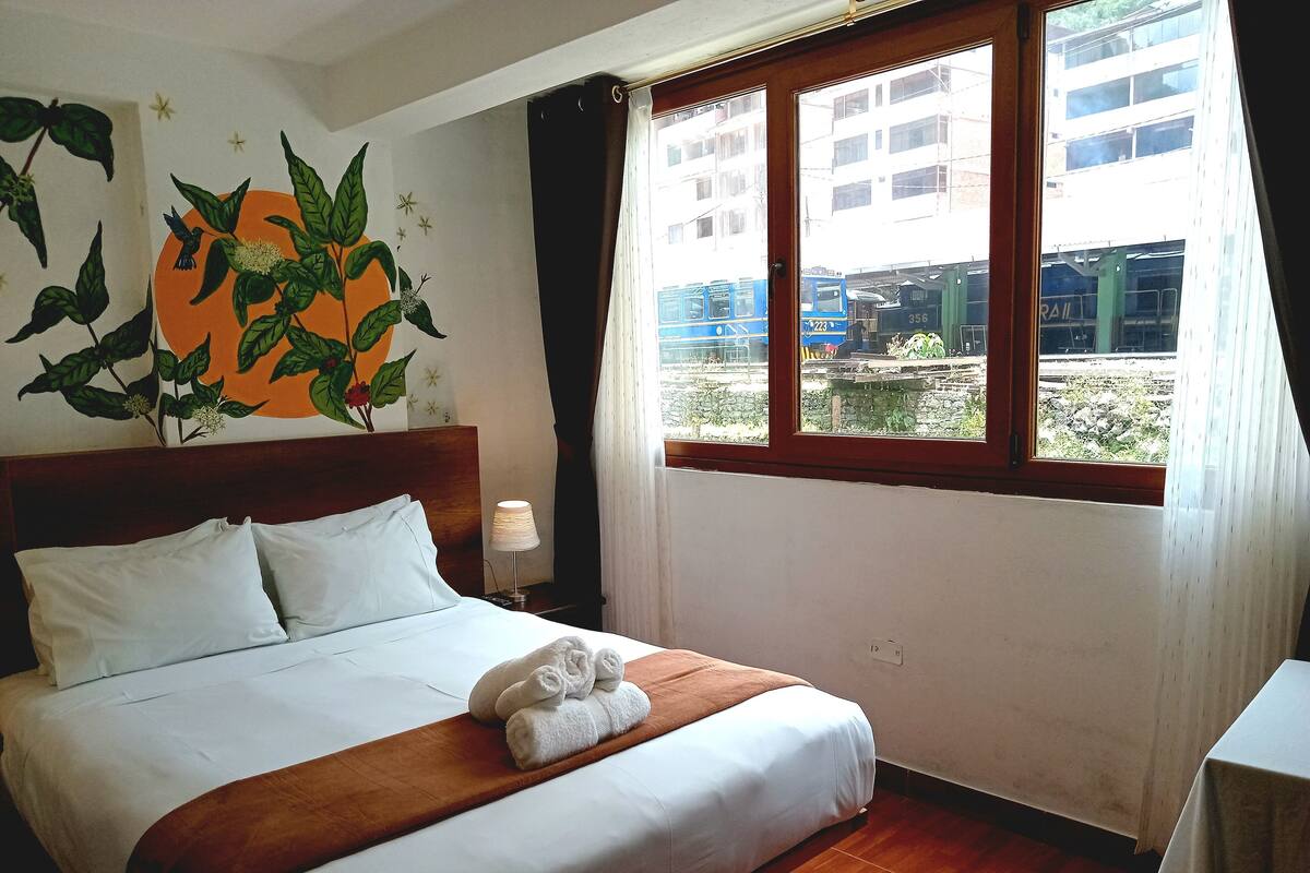 Ein Hotelzimmer mit einem Bett, einem Fenster mit Blick auf Gebäude und einen Zug, sowie einer Wand mit Pflanzen und Sterndekorationen.