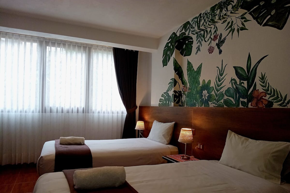 Ein Hotelzimmer mit zwei Betten, einem großen Fenster mit transparenten Vorhängen, einem Wandbild mit tropischen Blättern und einem Holzkopfbrett.