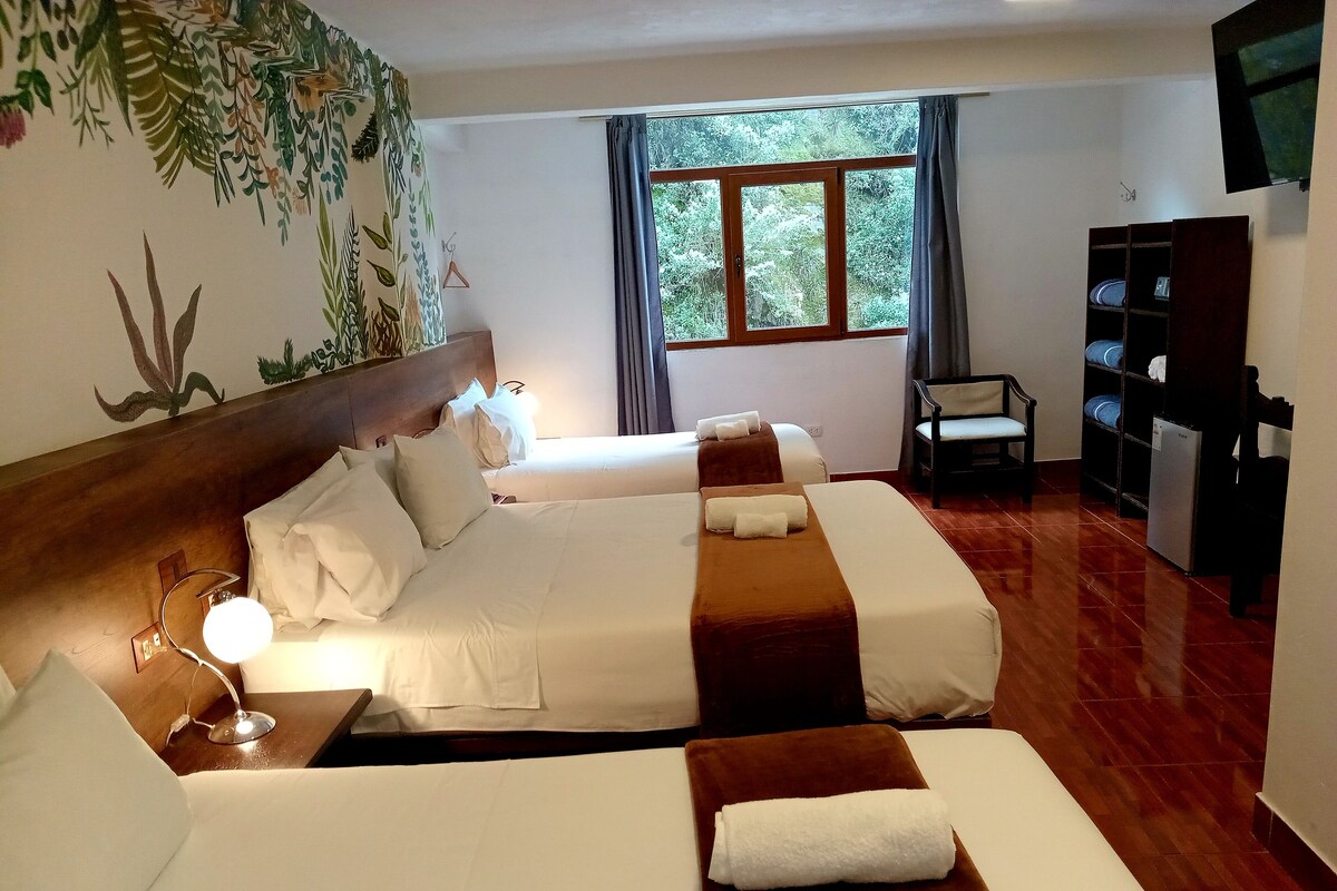 Ein Hotelzimmer mit zwei Betten, einem Holzkopfende, einem Nachttisch mit Lampe, einem Fenster mit Vorhängen, einem Sessel und einem an der Wand befestigten Fernseher.
