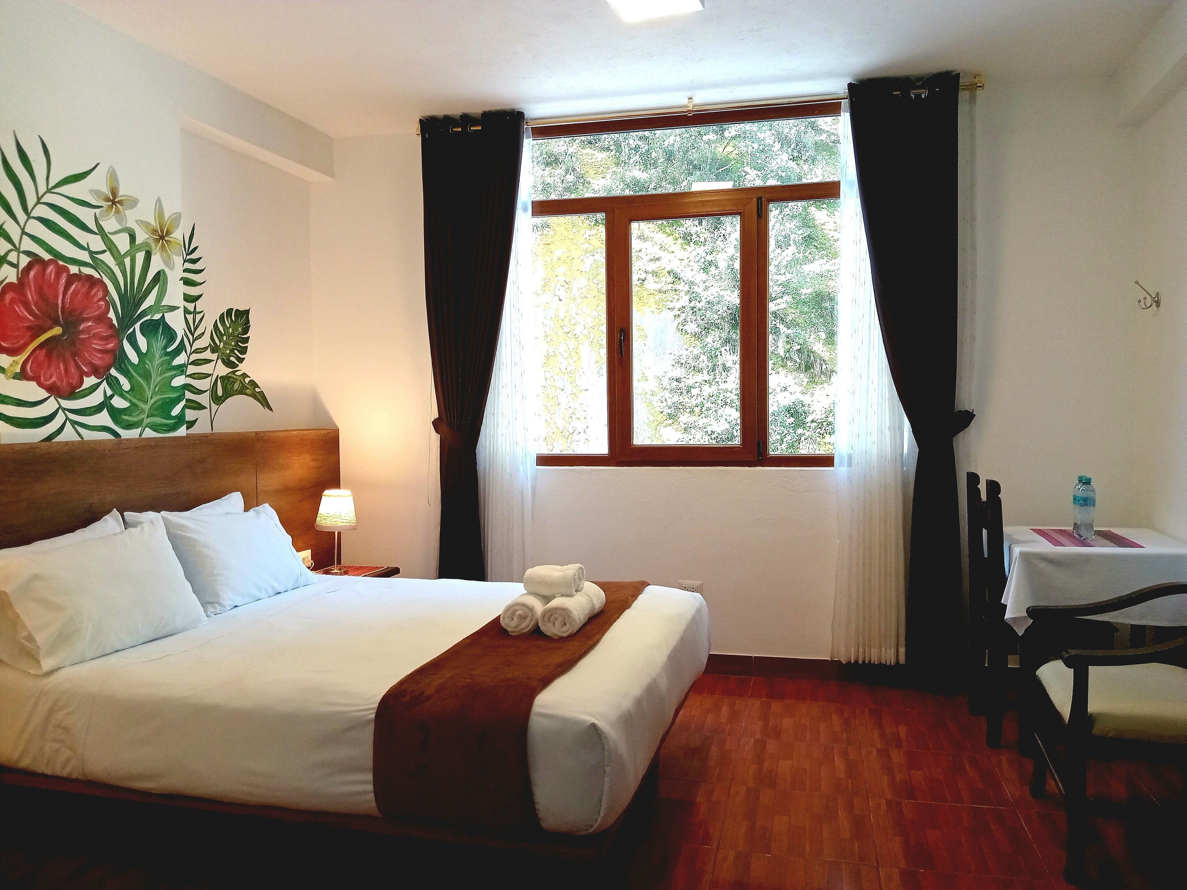 Quarto de hotel com cama, mesa de cabeceira, mesa com cadeira e janela com cortinas.
