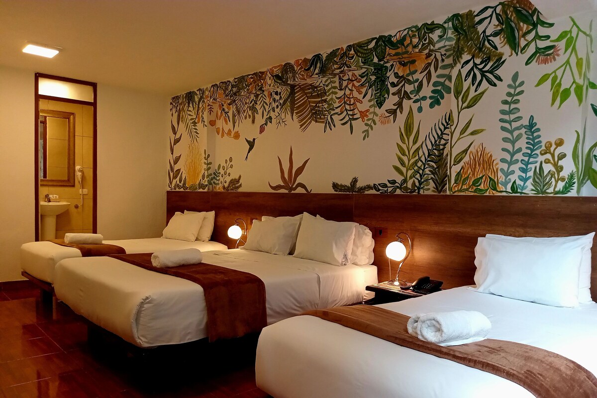 A hotel room with two beds, a wooden headboard, a wall mural, a bedside table with a lamp, and a bathroom with a sink and toilet.