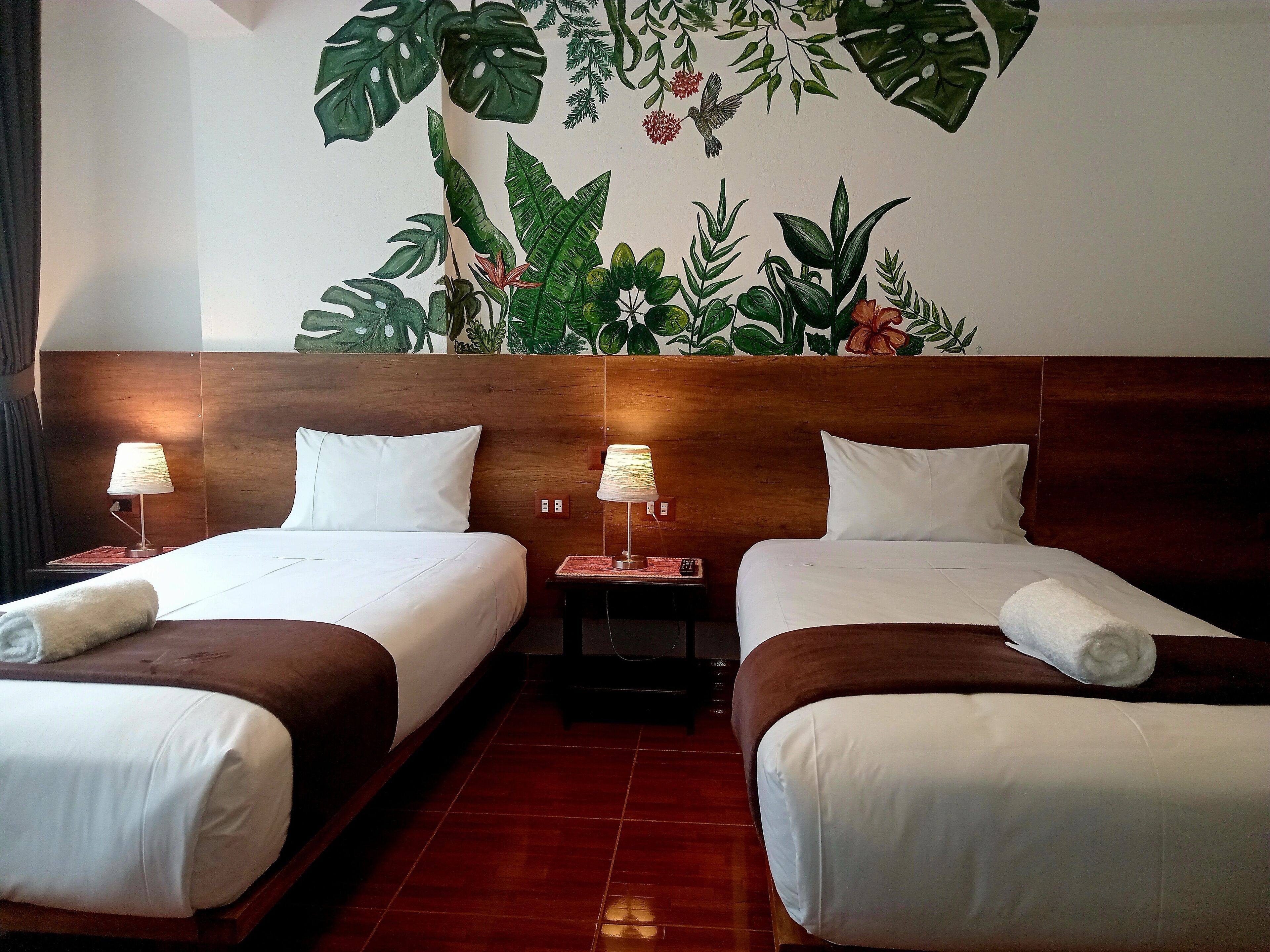 A hotel room with two beds, wooden headboard, white bedding, two bedside lamps, and a tropical-themed wall mural.