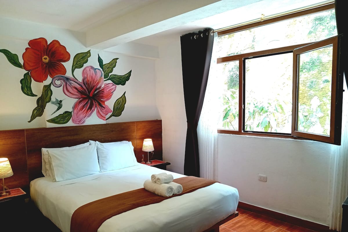 Ein Hotelzimmer mit Bett, Nachttisch, Lampe, Fenster mit Vorhängen und einer Wandmalerei mit floralem Muster.