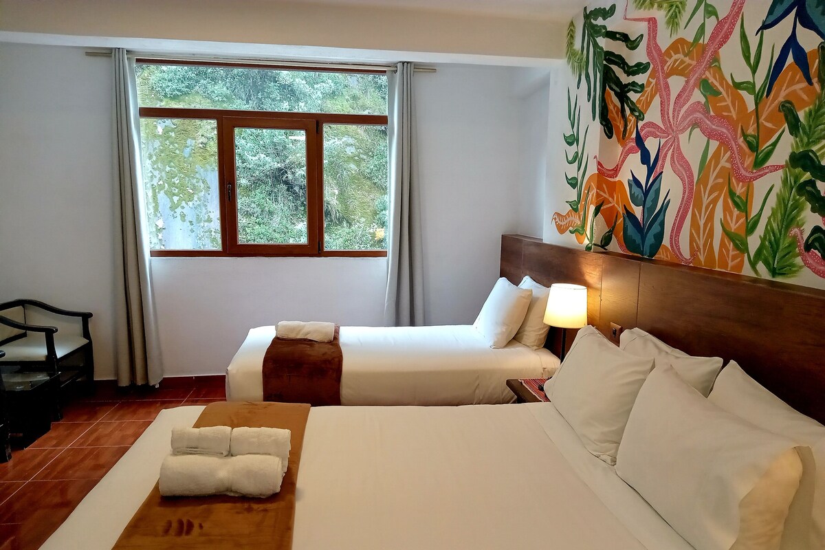 A hotel room with two beds, a wooden headboard, a large window with curtains, a desk, and a colorful wall mural.
