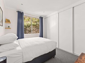 A bedroom with a bed, a window with curtains, a wardrobe, and a small plant on a shelf. - Beautifully Peaceful 2-bed Apartment (Sydney)
