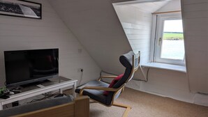 Een kamer met een schuine plafond, een televisie op een wit standaard, een houten schommelstoel en een raam met uitzicht op zee.