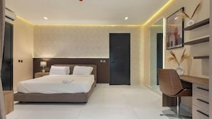 A modern hotel room with a large bed, a desk, and a chair. There is a door and a wall-mounted shelf with decorative items. - Chatkrum Hotel (Abuja)