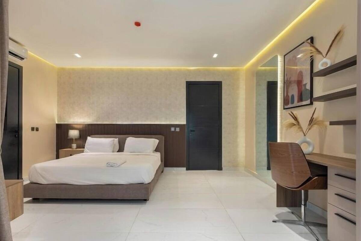 A modern hotel room with a large bed, a desk, and a chair. There is a door and a wall-mounted shelf with decorative items.