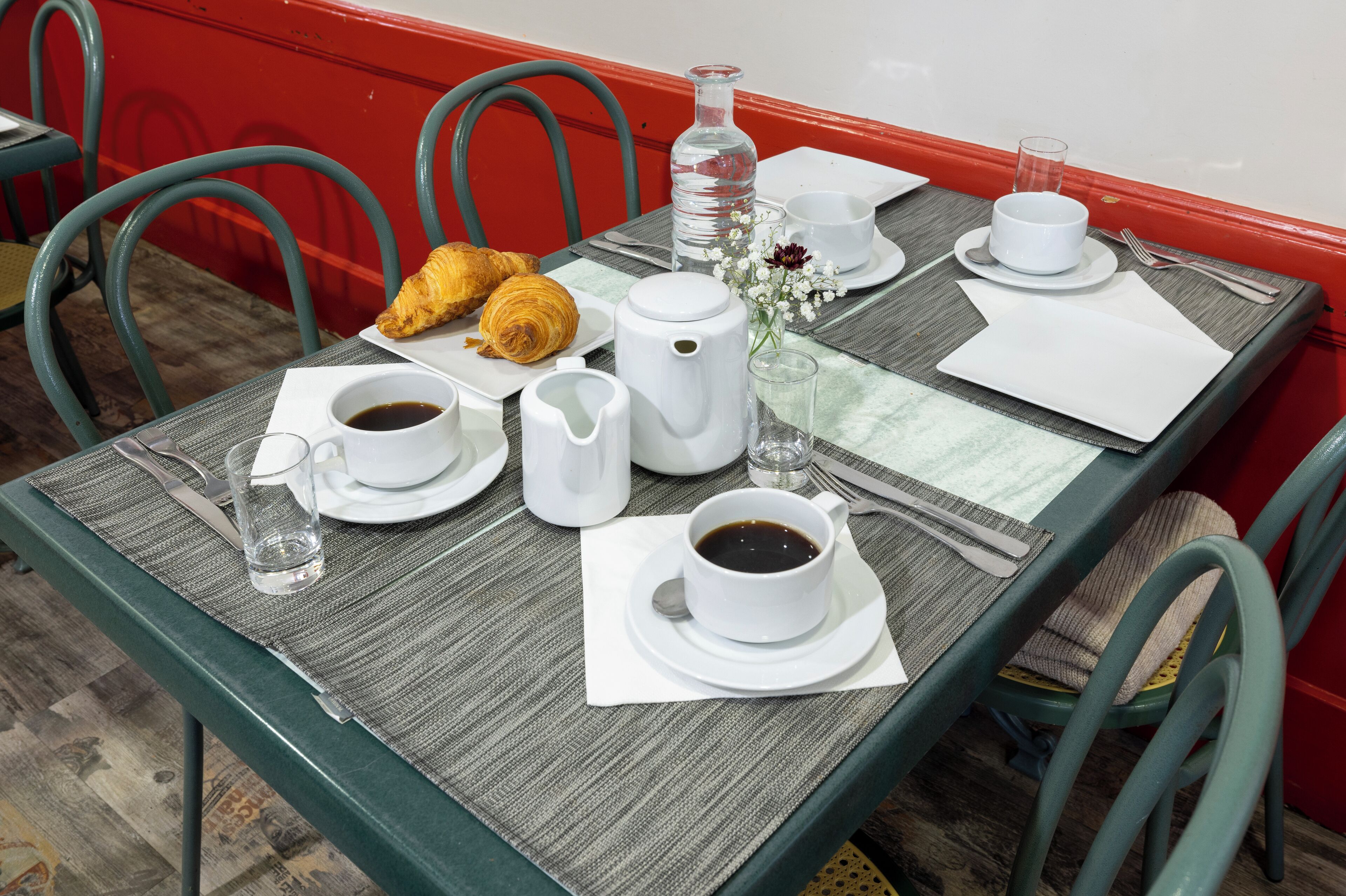 Una mesa de desayuno servida con croissants, tazas de café, una botella de agua y un florero con flores.