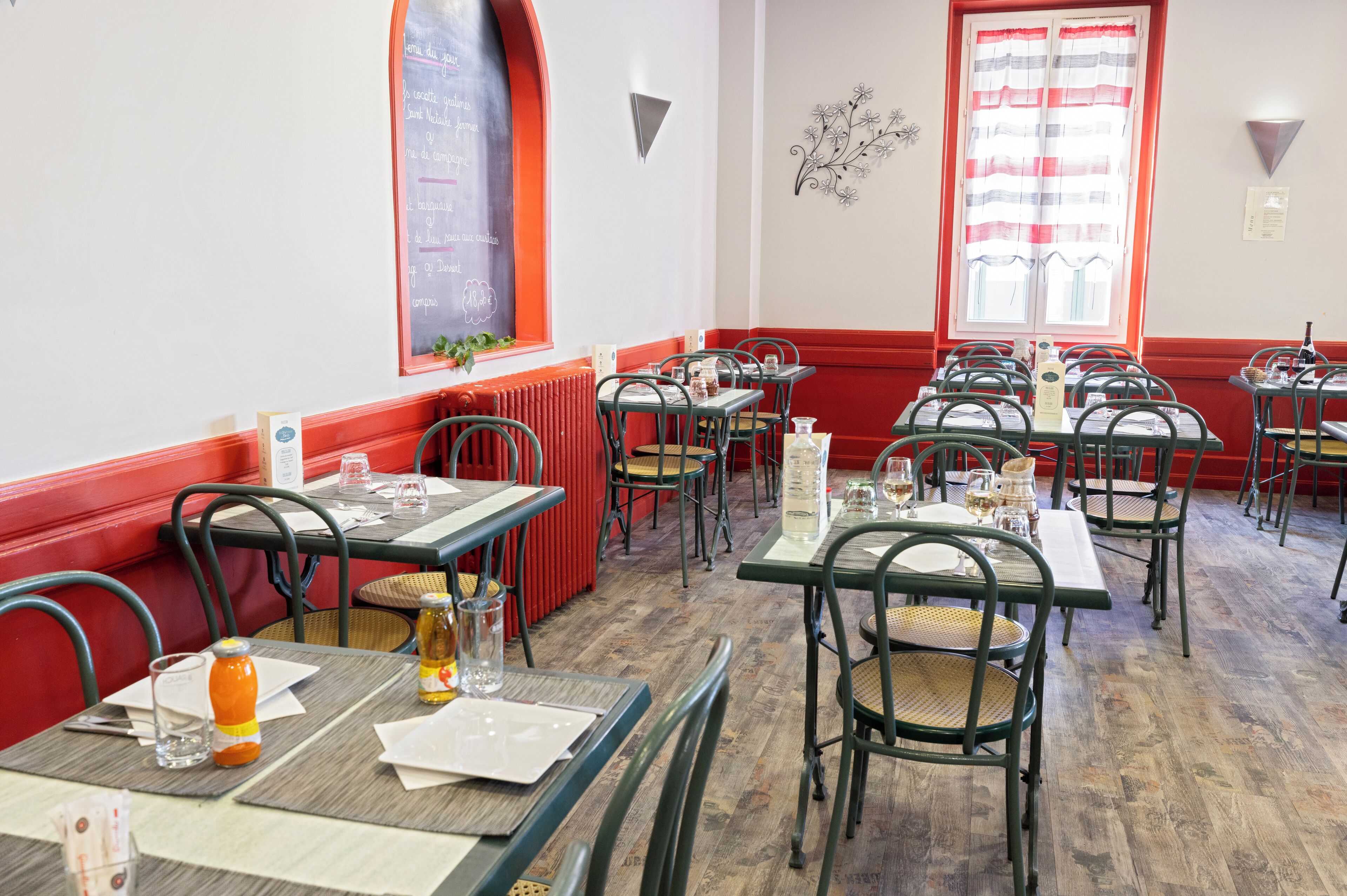 L’intérieur d’un restaurant, avec de nombreuses tables et chaises, un menu écrit à la craie et une fenêtre ornée de rideaux.