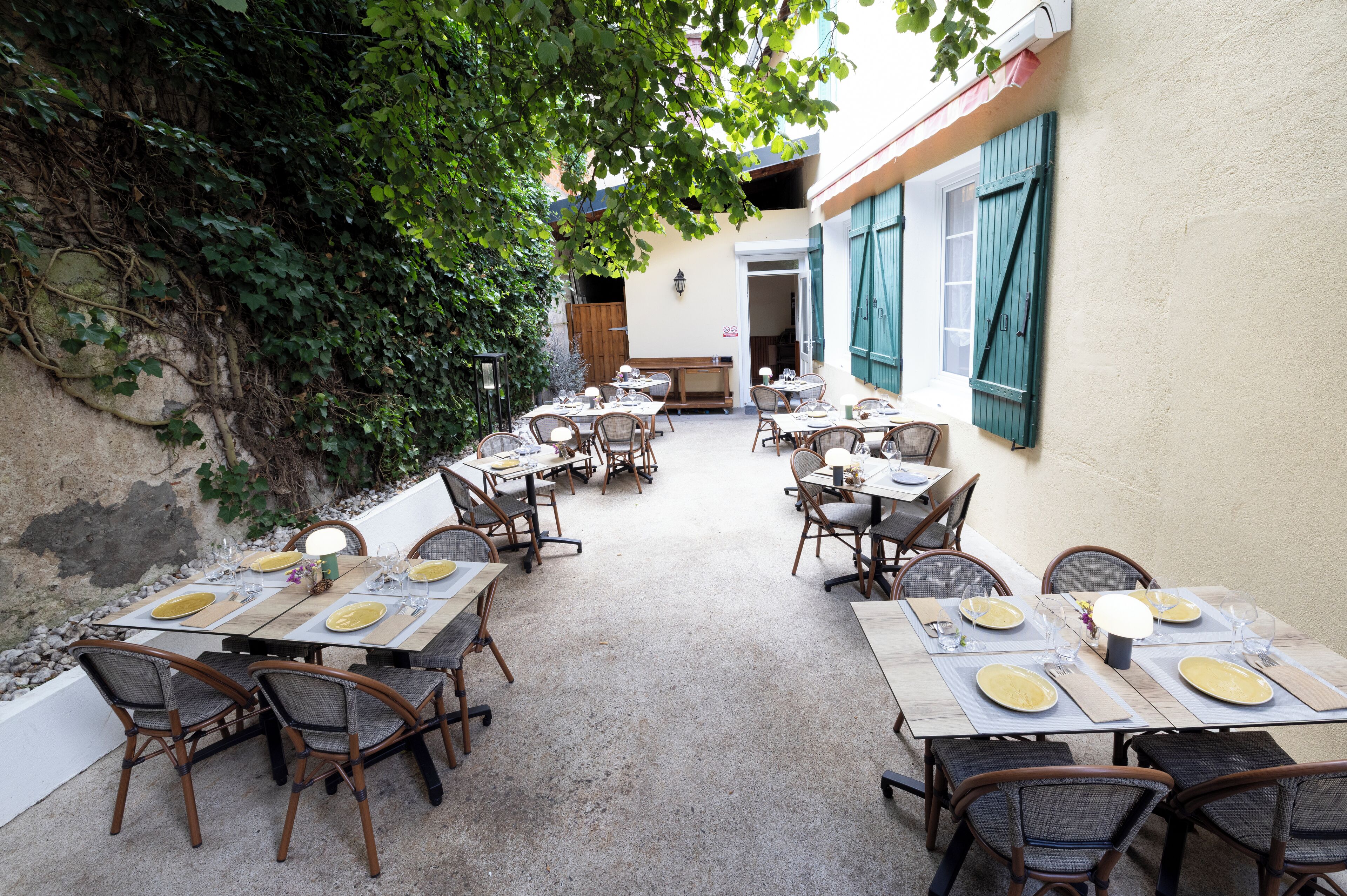 An outdoor dining area with tables and chairs, surrounded by a stone wall and greenery.