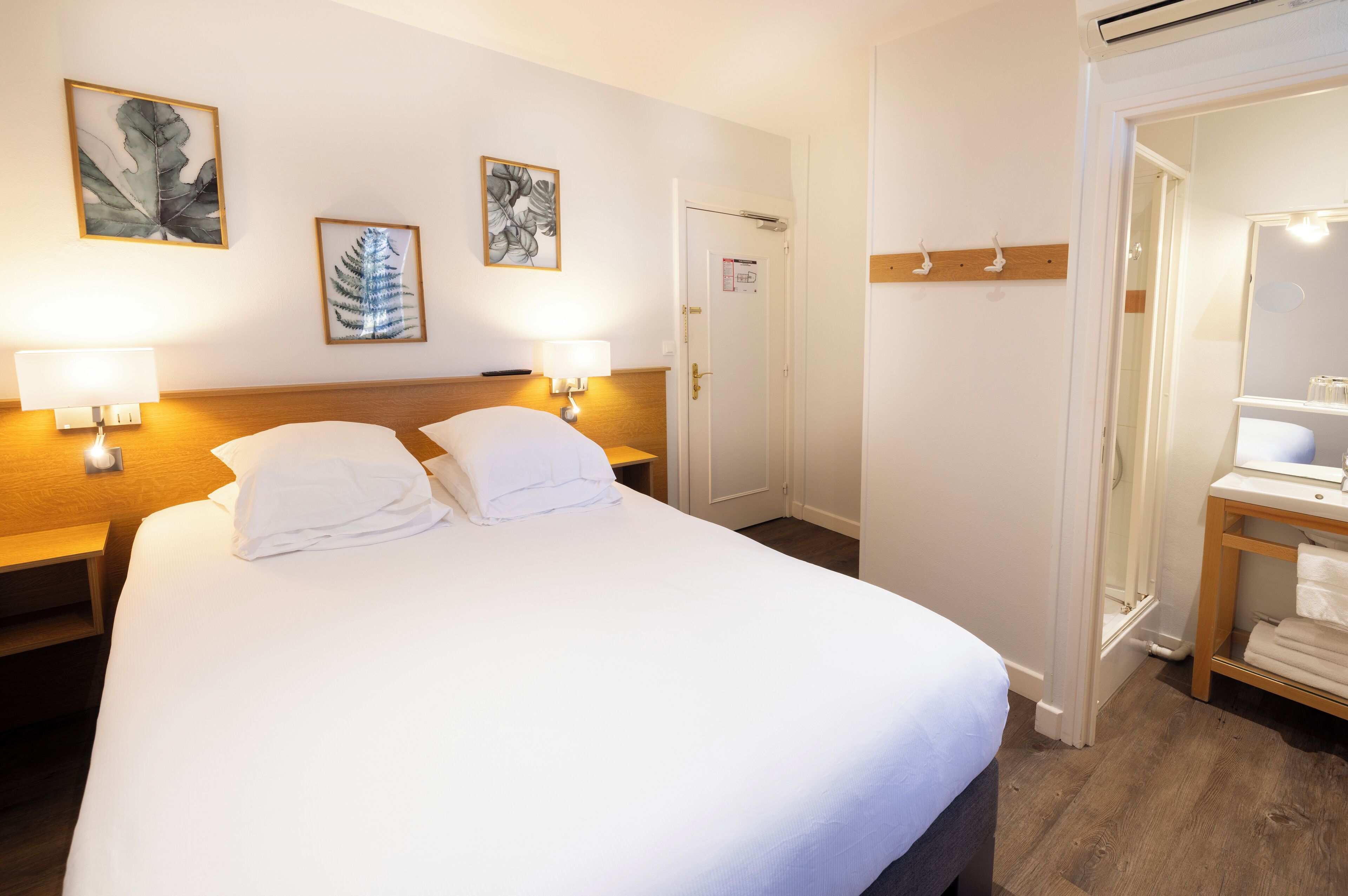 A hotel room with a large bed, two bedside tables with lamps, a wooden headboard, and two framed leaf artworks on the wall.