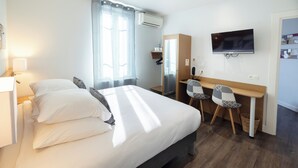 A hotel room with a bed, a desk with chairs, a television, and an air conditioning unit. - Hotel Restaurant de Paris (Jaligny-sur-Besbre)