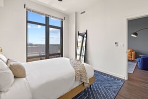 A modern bedroom with a large bed, a view of the city through floor-to-ceiling windows, and a small seating area with a lamp. - 4 Units | Arlington Haven | Pet & Family Friendly | Near DC Monuments & Trails (Arlington)