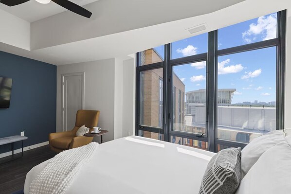 A modern hotel room with a large bed, a chair, and a view of the city through large windows.