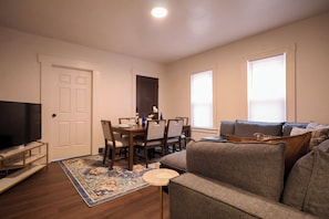 A living room with a sofa, a TV, a dining table with chairs, and a rug. - The Weyl Collective 6BR Professional Retreat Near Downtown Rochester (Rochester)