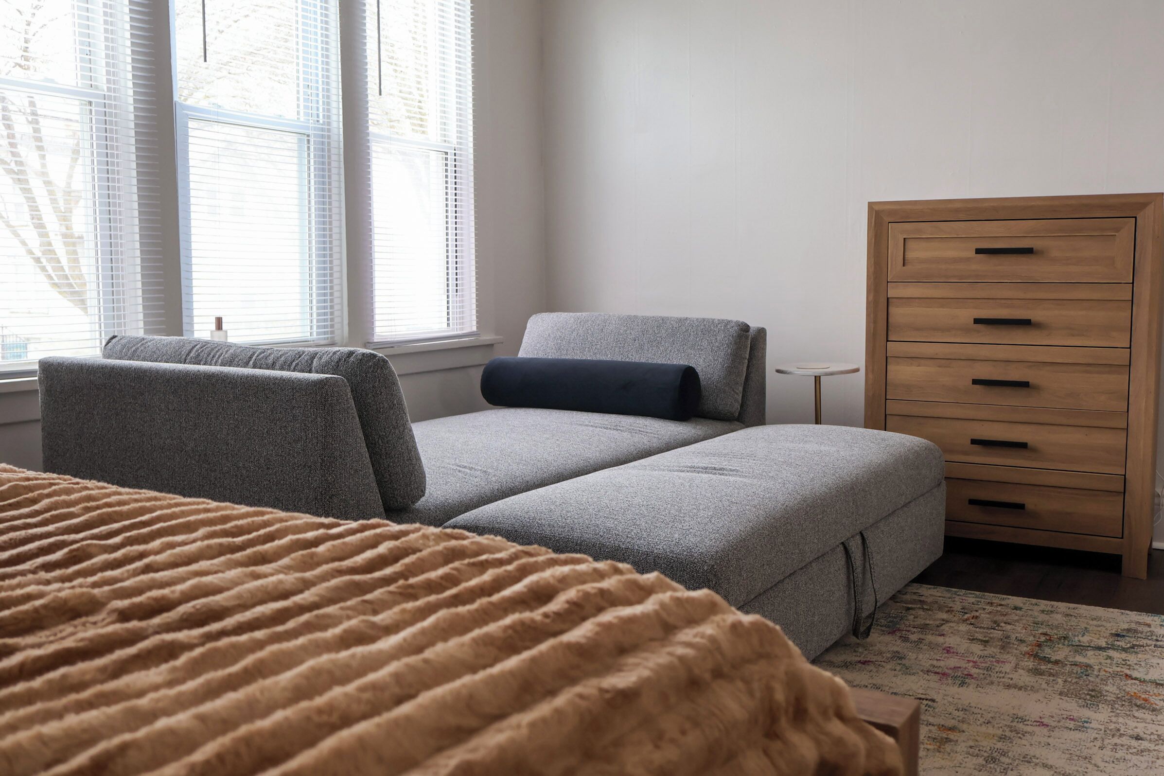 A bedroom with a bed, a grey sofa, a wooden dresser, and a window with blinds.