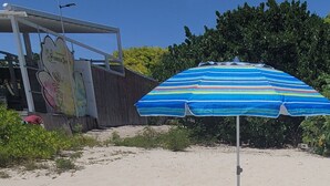 A beach with two blue lounge chairs under a striped umbrella, a Ferrero sign, and a fence with greenery.