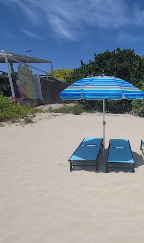 Una playa con dos sillas azules para tomar el sol bajo una sombrilla a rayas, un letrero de Ferrero y una valla con follaje.