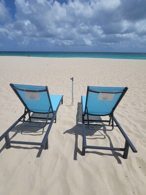 Two blue lounge chairs on a sandy beach with a clear sky and ocean in the background.