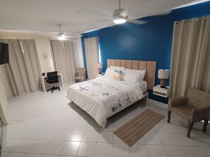 A hotel room with a bed, two chairs, a desk, a ceiling fan, and a TV.