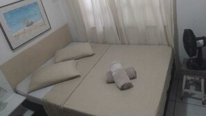 En snyggt bäddad säng med ett randigt överkast och två kuddar, ett litet sängbord med en fläkt och en inramad tavla på väggen.
